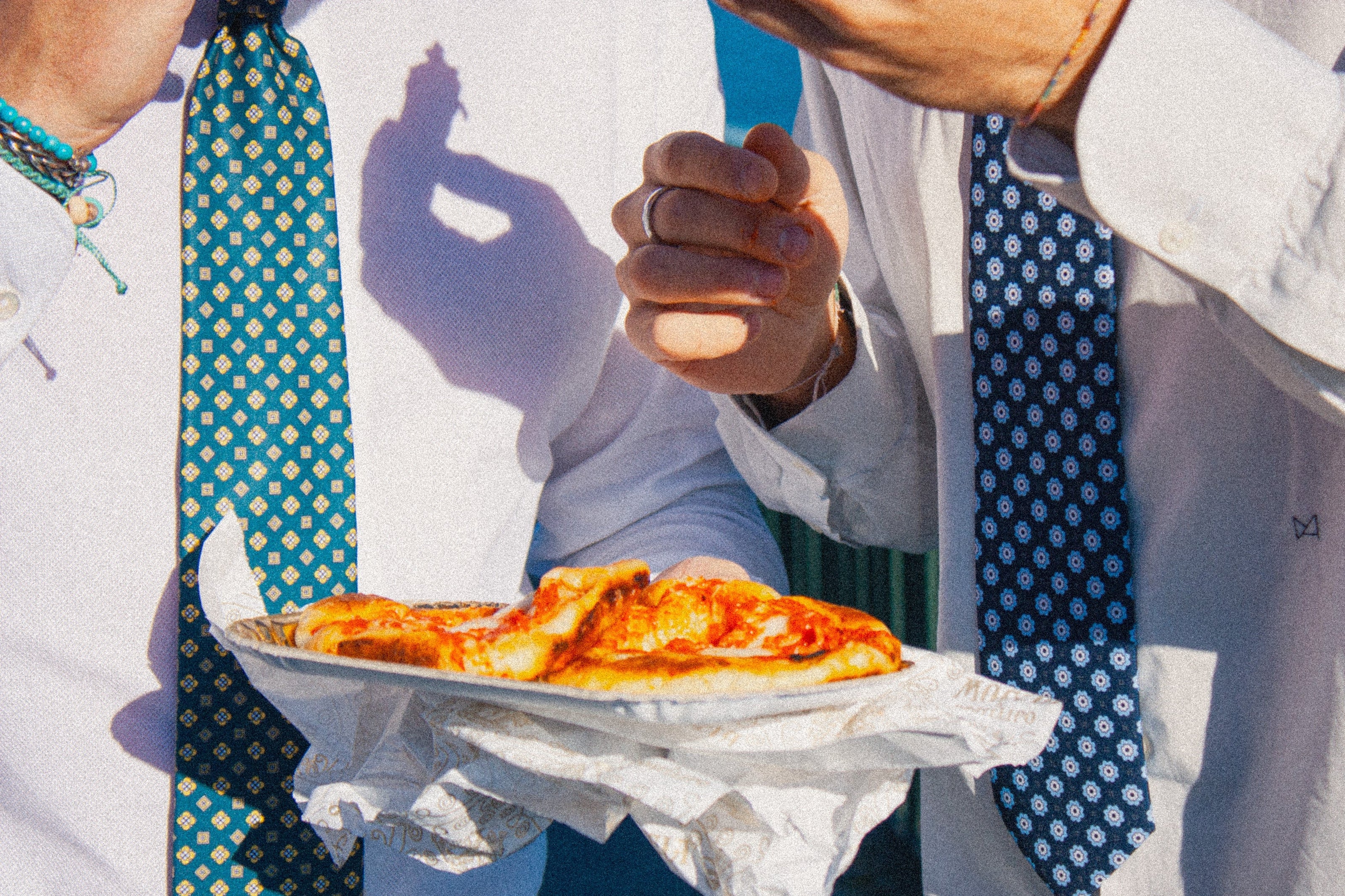 Due uomini in pausa pranzo con camicia e cravatta TieTiè che mangiano delle pizzette di Moccia con le mani a Via Petrarca a Posillipo, Napoli davanti al Serpentone. Le cravatte sono verdi e blu.